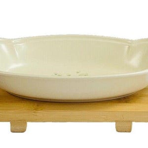 May include: A white ceramic serving bowl with two handles, sitting on a wooden stand with legs. The bowl has a slightly oval shape and a smooth, glossy finish.
