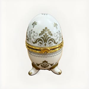 May include: A white porcelain egg-shaped trinket box featuring gold-colored filigree patterns and floral motifs. The container has a hinged center with a gold-colored metal rim and clasp, supported by three small, curved porcelain feet with gold tips.