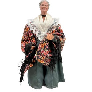 May include: A detailed figurine of an elderly woman in traditional attire. She wears a black shawl with a floral pattern, a white lace collar, and a dark green skirt. The figurine has a realistic painted face and gray hair.