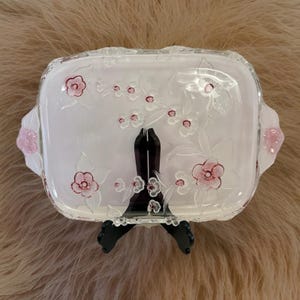 May include: A rectangular glass serving tray with a white interior and clear, floral-patterned exterior. The tray features pink flowers and leaves, with pink accents on the handles. It is displayed on a black stand.