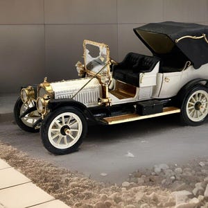 May include: A white vintage car with a black canvas top and gold accents. The car has a wooden dashboard and a spare tire on the back.