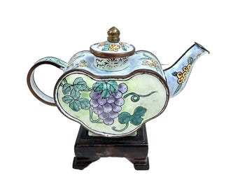 Puede incluir: Una tetera de cloisonné azul y verde con un diseño de vid. La tetera tiene una tapa y un pico y se asienta sobre un soporte de madera.