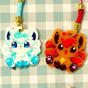 May include: Two cartoon fox charms with lanyards. One is white and blue, the other is orange and red. Both have gold-colored metal accents. The charms are on a light blue and white checkered background.