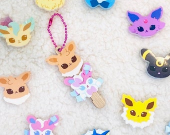 May include: A selection of colourful character-shaped charms, including Eevee and its evolutions, in various poses. One charm is attached to a pink beaded chain. The charms are in shades of brown, blue, yellow, purple, and pink.