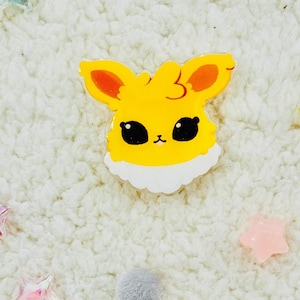 Puede incluir: Un personaje de dibujos animados amarillo y naranja con ojos grandes y un cuello blanco esponjoso. El personaje es un pin hecho de acr&iacute;lico transparente.