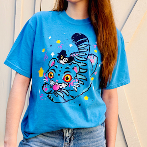 May include: A light blue t-shirt featuring a cartoon tiger with a black hat and a striped tail. The tiger has large yellow eyes, a pink tongue, and is surrounded by stars and confetti. The shirt is made of a soft material.