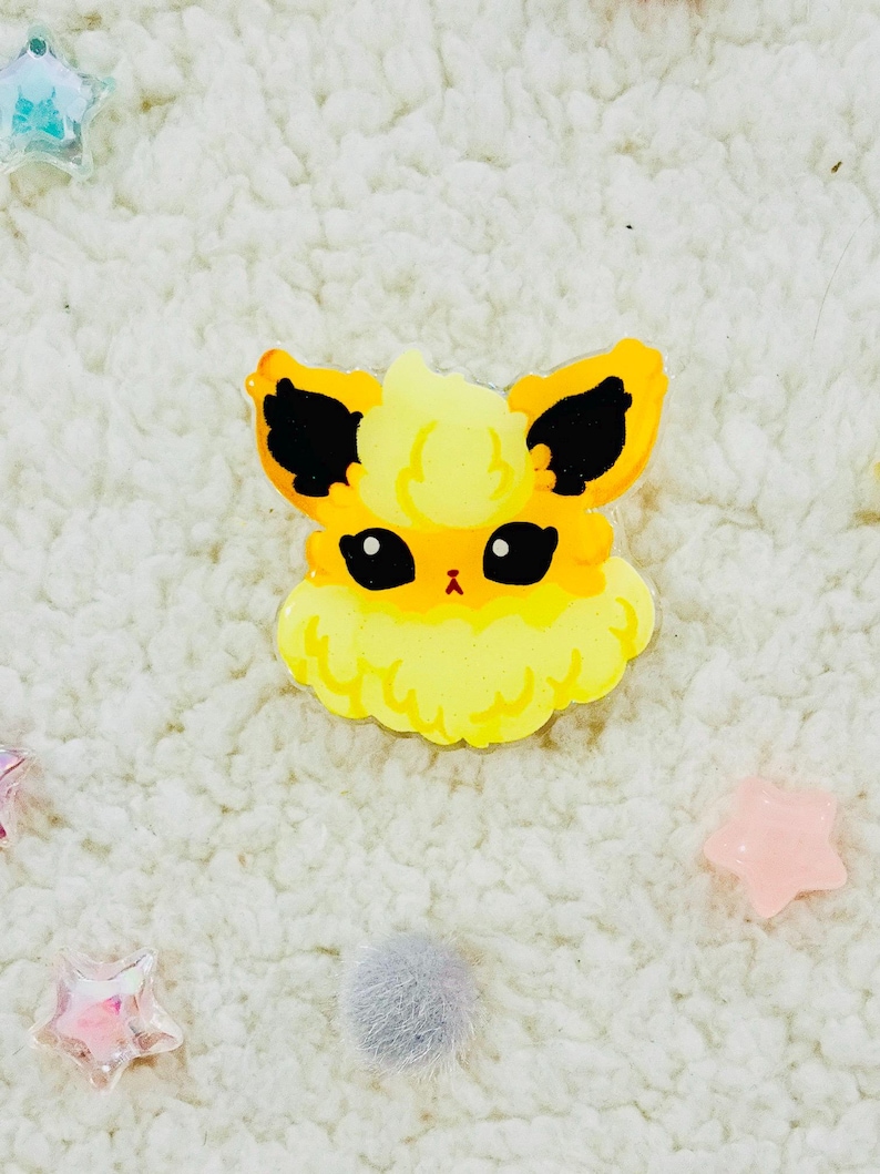 Puede incluir: Un personaje de dibujos animados amarillo y negro con pelaje esponjoso y ojos grandes. El personaje es un pin hecho de acr&iacute;lico transparente.