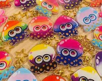 Peut inclure: Un ensemble de porte-clés translucides et colorés représentant une pieuvre de dessin animé aux grands yeux. Chaque porte-clés présente un dégradé de couleurs différent et est décoré de paillettes. Les porte-clés sont attachés à des breloques en forme d'étoile dorée.