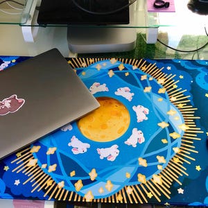 May include: A blue and gold mousepad with a moon and stars design. The mousepad features a circular design with a large yellow moon in the center, surrounded by white bunnies and stars. The mousepad is on a desk with a laptop and a pink mouse.
