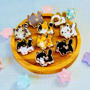 May include: A collection of cat-shaped enamel pins in various colors, including gray, calico, orange, and black, with gold-colored outlines. The pins are arranged on a round wooden tray, surrounded by star-shaped and other decorative elements.
