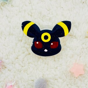Puede incluir: Un amuleto de resina negra y amarilla con purpurina de un personaje de Pokemon con ojos rojos. El personaje tiene un solo c&iacute;rculo amarillo en la frente.