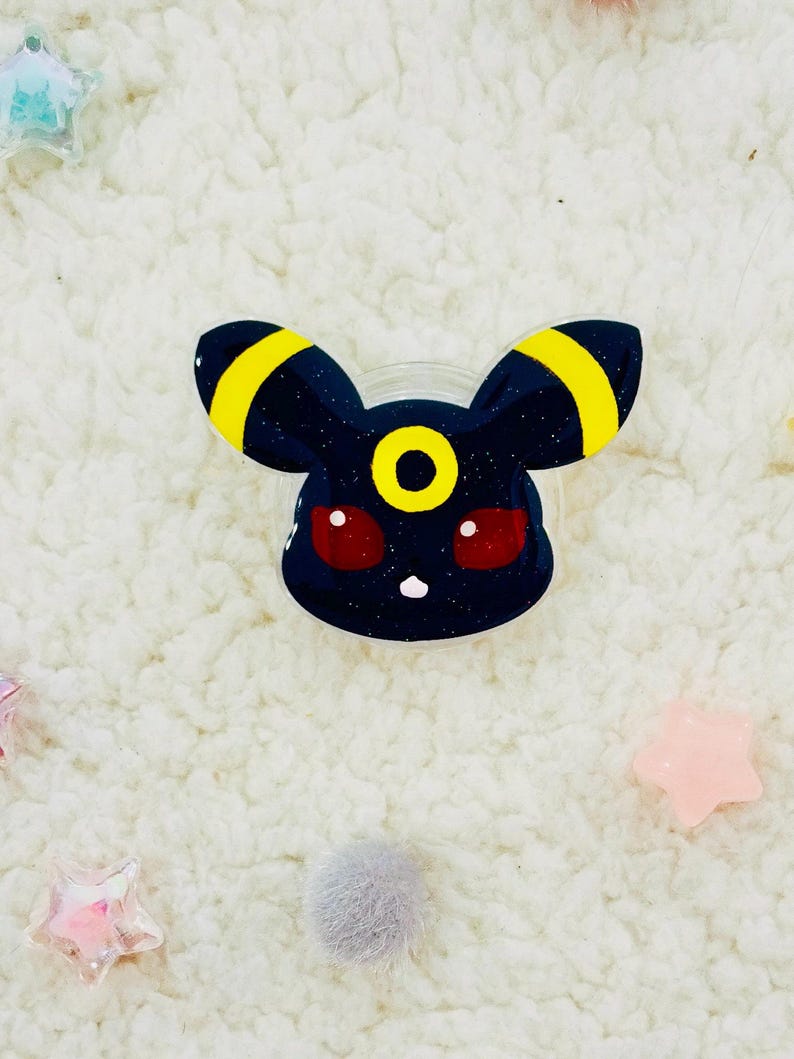 Puede incluir: Un amuleto de resina negra y amarilla con purpurina de un personaje de Pokemon con ojos rojos. El personaje tiene un solo c&iacute;rculo amarillo en la frente.