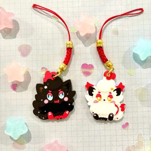 May include: Two black and white cartoon character charms with red accents on red string phone charms. The charms are attached to red string phone straps with gold beads.