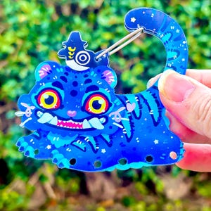 Puede incluir: Un llavero azul con forma de gato con un diseño caprichoso. El llavero presenta un gato de dibujos animados con grandes ojos amarillos, una boca rosa y una corona decorativa. El cuerpo del gato está adornado con estrellas y patrones blancos. Un cierre de metal plateado está unido al llavero.