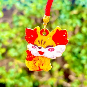 May include: A yellow and red fox charm with a red cord. The fox has white accents, a smiling face, and star details. The charm is hanging against a blurred green background.