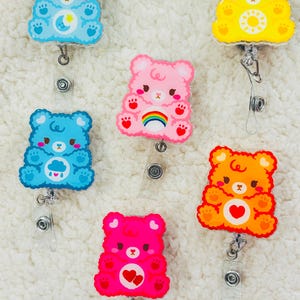 May include: Six colorful cartoon bear badge holders with different designs. The bears are blue, pink, yellow, orange, and red. Each bear has a different design on its chest, including a moon and star, a rainbow, a sun, a heart, and a cloud with raindrops.