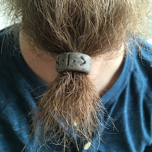 May include: A gray beard bead with two runes carved into it. The bead is tied around a long, brown beard.