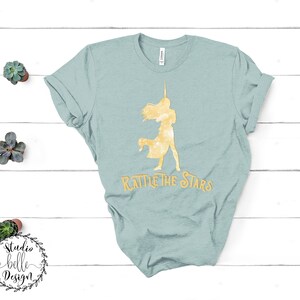 Puede incluir: Una camiseta azul claro con una silueta dorada de una mujer sosteniendo una estrella y el texto "Rattle the Stars".