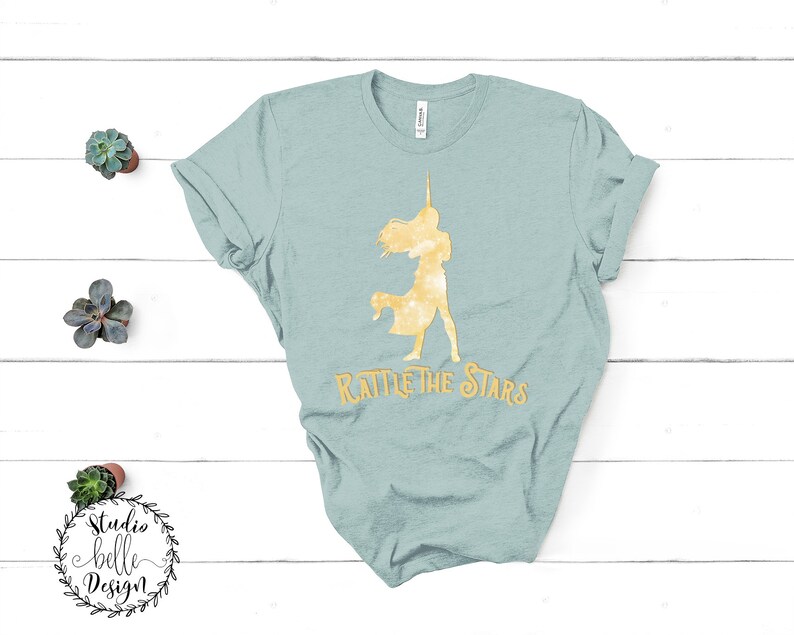 Puede incluir: Una camiseta azul claro con una silueta dorada de una mujer sosteniendo una estrella y el texto "Rattle the Stars".