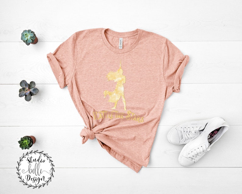 Puede incluir: Una camiseta de color melocot&oacute;n con una silueta dorada de una mujer sosteniendo una espada y el texto "To the Stars".