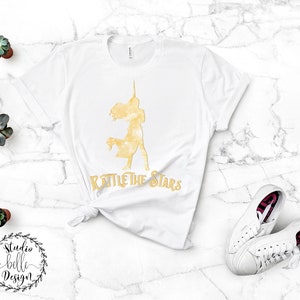 Puede incluir: Camiseta blanca con una silueta dorada de una mujer con un cuerno de unicornio y el texto "Rattle the Stars".