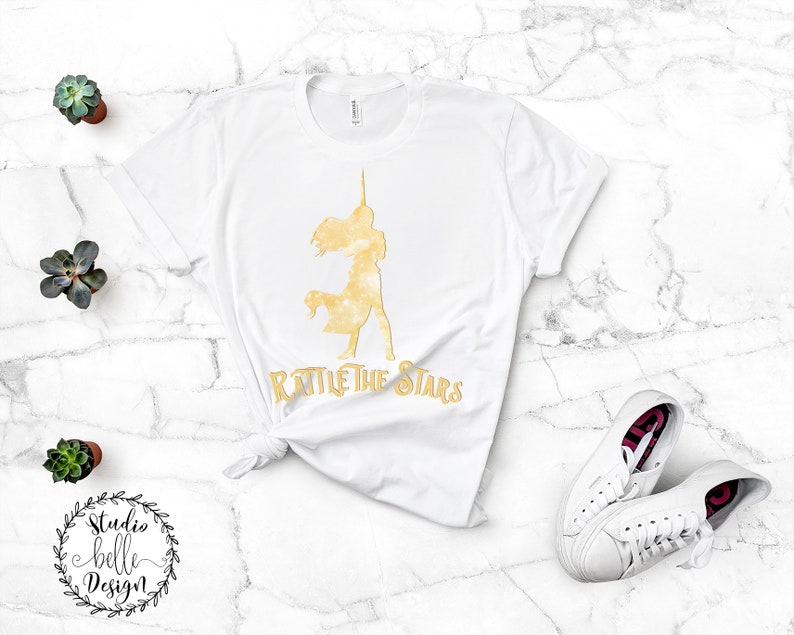 Puede incluir: Camiseta blanca con una silueta dorada de una mujer con un cuerno de unicornio y el texto "Rattle the Stars".