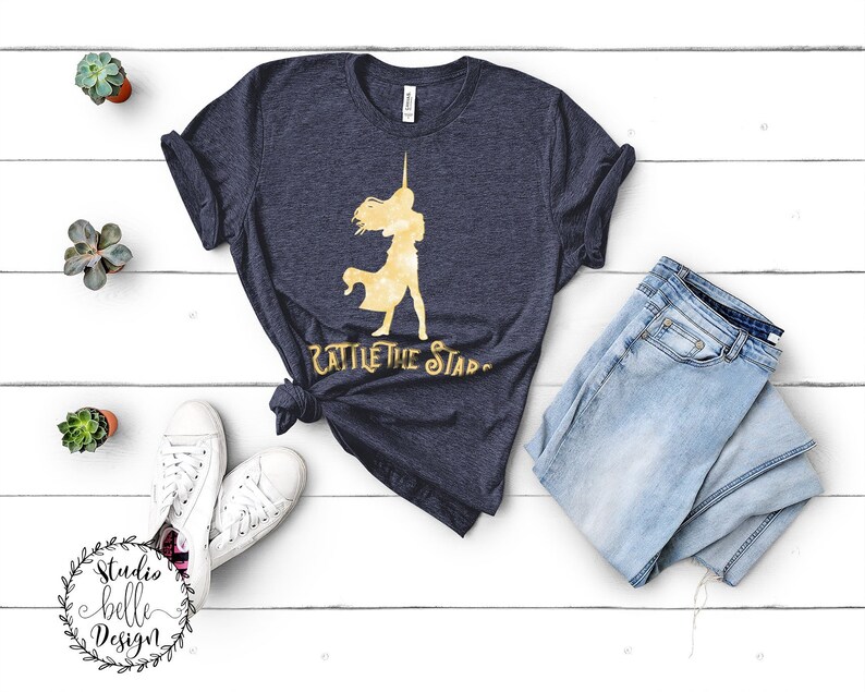 Puede incluir: Una camiseta azul marino con una silueta dorada de una mujer sosteniendo una espada y el texto "Battle the Stars".