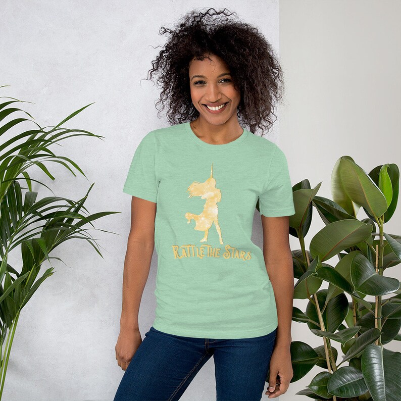 Puede incluir: Una camiseta verde claro con una silueta dorada de una mujer sosteniendo una espada y el texto "Rattle the Stars".