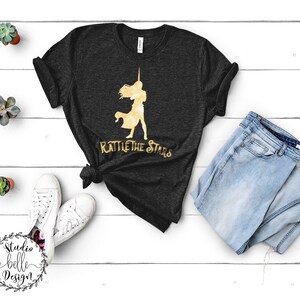 Puede incluir: Una camiseta negra con una silueta dorada de una mujer sosteniendo una espada y el texto "Rattle the Stars".