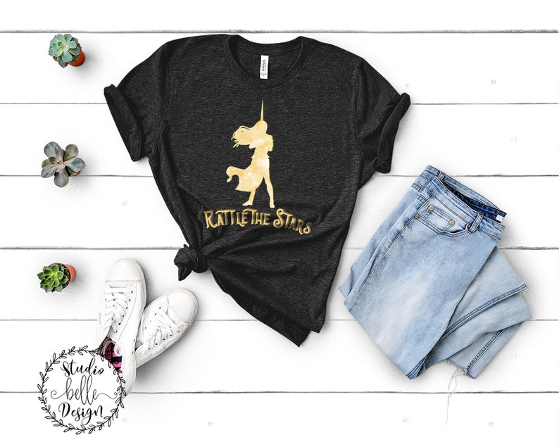 Puede incluir: Una camiseta negra con una silueta dorada de una mujer sosteniendo una espada y el texto "Rattle the Stars".