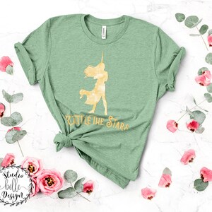 Puede incluir: Una camiseta verde con una silueta dorada de una mujer sosteniendo una espada y el texto "Battle the Stars".