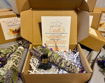May include: An open cardboard box containing a purification kit. Inside, a selenite and black tourmaline crystal cluster, anointing oil, and a smudging stick are visible. The box also includes a card with the text "House Blessing Purification Kit".