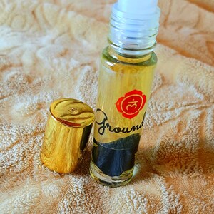 May include: A clear glass roller bottle with a gold cap and a red and white label that says "Ground" and a symbol. The bottle is filled with a yellow liquid.