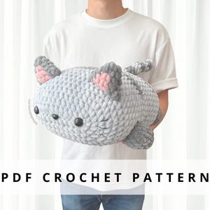 以下が含まれることがあります： ピンク色の内耳と黒いボタンの目を持つ、大きな灰色の編み猫のぬいぐるみ。白いTシャツを着た人がぬいぐるみを持っています。下部のテキストは「PDF CROCHET PATTERN」と書かれています。