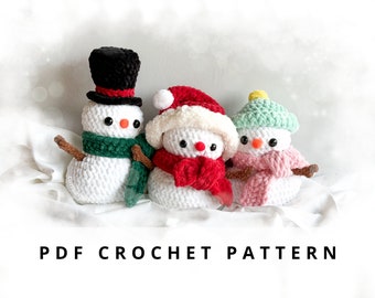 May include: Three crocheted snowman amigurumi toys. The snowman on the left is wearing a black top hat, a green scarf, and a brown twig nose. The snowman in the middle is wearing a red and white Santa hat and a red scarf. The snowman on the right is wearing a green hat and a pink scarf.