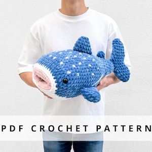 Könnte beinhalten: Ein großer, blau gehäkelter Wal-Plüsch mit weißen Punkten, Flossen und einem rosa-weißen Maul. Der Wal wird vor einer Person gehalten, die ein weißes T-Shirt trägt. Der Text auf dem Bild lautet "PDF CROCHET PATTERN".
