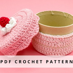 May include: A pink crocheted cupcake-shaped box with a white crocheted lid and a red strawberry on top. The box is open and the inside is lined with brown paper. The text "PDF CROCHET PATTERN" is printed below the box.