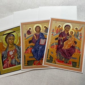 May include: Three religious icon cards featuring traditional Byzantine art. The first card depicts a seated angel with wings, holding a white flower. The second card shows a seated figure with a book, and the third card shows a mother and child with two angels above them.