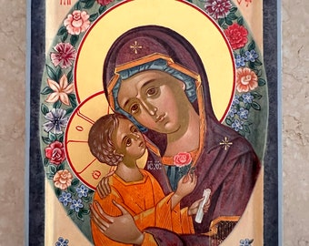 Könnte beinhalten: Eine religiöse Ikone, die die Jungfrau Maria zeigt, die ein Jesuskind hält. Die Ikone ist auf einem Holzpaneel gemalt und in einem dunkelblauen Rahmen gerahmt. Die Jungfrau Maria trägt eine blaue Robe und einen roten Schal. Sie hält das Jesuskind in ihren Armen. Das Jesuskind trägt eine weiße Robe. Die Ikone ist von einem Kranz aus Blumen umgeben.