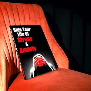 May include: A black book titled "Ride Your Life Of Stress & Anxiety" sits on an orange velvet chair. The book's cover has white and red text, with an illustration of a person's face and hands. The chair has a vertical ribbed design.