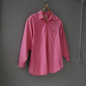 May include: A pink long-sleeved button-down shirt with a small pocket on the left side. The shirt is hanging on a wooden hanger against a gray wall.