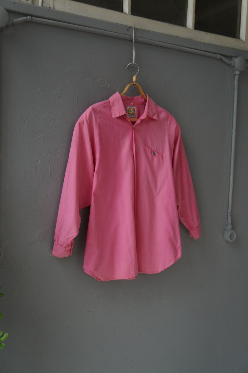 May include: A pink long-sleeved button-down shirt with a small pocket on the left side. The shirt is hanging on a wooden hanger against a gray wall.