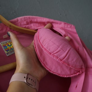 May include: A pink button-up shirt with a sewn-on patch that says "California Surfwear" in rainbow lettering. The shirt is hanging on a wooden hanger.