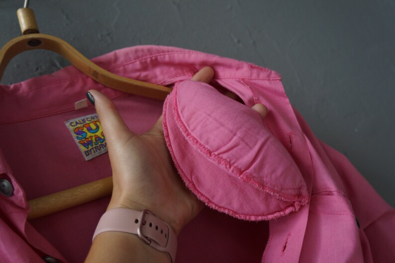 May include: A pink button-up shirt with a sewn-on patch that says "California Surfwear" in rainbow lettering. The shirt is hanging on a wooden hanger.