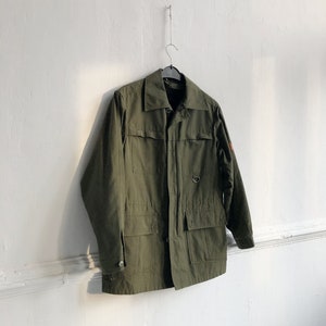 May include: A green utility jacket with a button closure, two large flap pockets, and a small pocket on the left chest. The jacket is hanging on a hanger against a white wall.