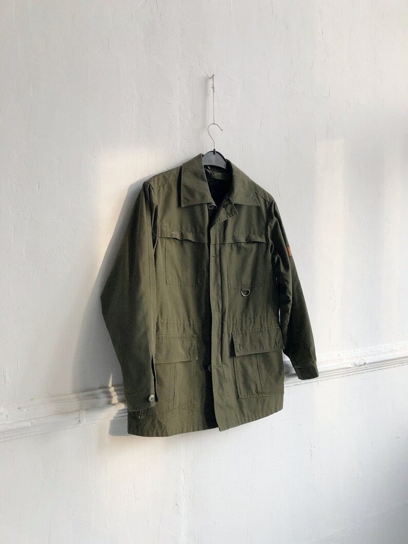 May include: A green utility jacket with a button closure, two large flap pockets, and a small pocket on the left chest. The jacket is hanging on a hanger against a white wall.