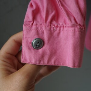 May include: Close-up of a pink button on a pink shirt cuff. The button is metal and has a single hole.