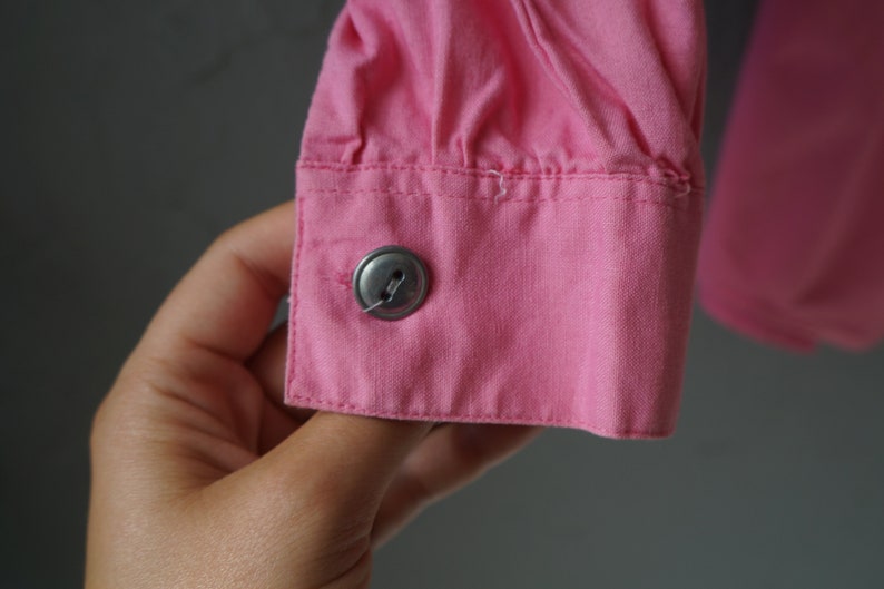 May include: Close-up of a pink button on a pink shirt cuff. The button is metal and has a single hole.