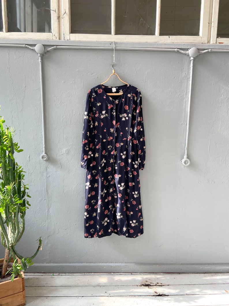 K&ouml;nnte beinhalten: Ein lang&auml;rmeliges, marineblaues Kleid mit einem Blumendruck mit rosa und wei&szlig;en Blumen. Das Kleid hat einen Knopfleisten vorne und eine lockere Passform.