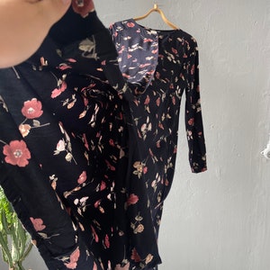 K&ouml;nnte beinhalten: Ein schwarzes Maxikleid mit Blumenmuster mit rosa und wei&szlig;en Bl&uuml;ten. Das Kleid hat lange &Auml;rmel und einen Knopfleisten vorne.